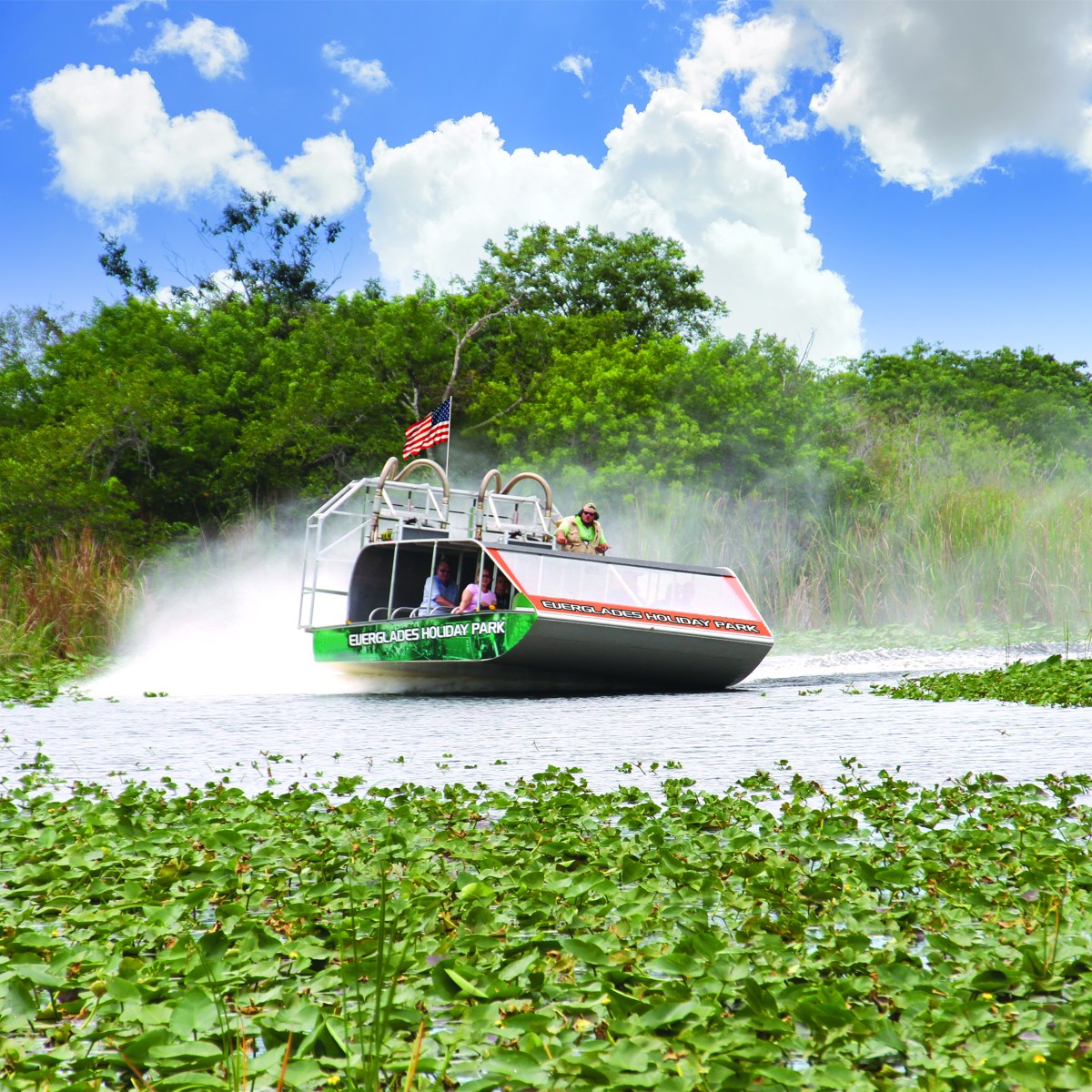 Airboat speeding through a lush, plant-filled marsh under a blue sky with clouds.