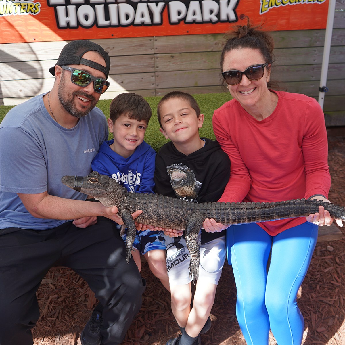 Four people sitting, holding a small alligator at a park.