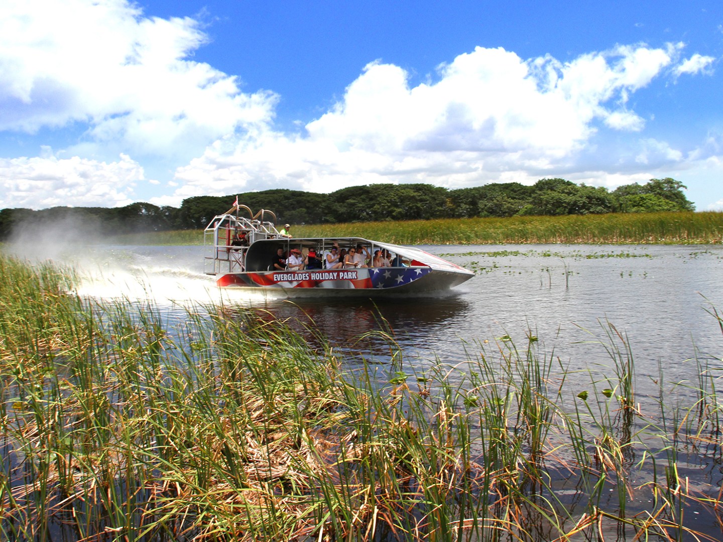 Airboat with people in wetland under blue sky with clouds.